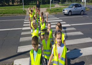 dzieci ubrane w kamizelki odblaskowe w kolorze cytrynowym przechodzą przez jezdnię po pasach